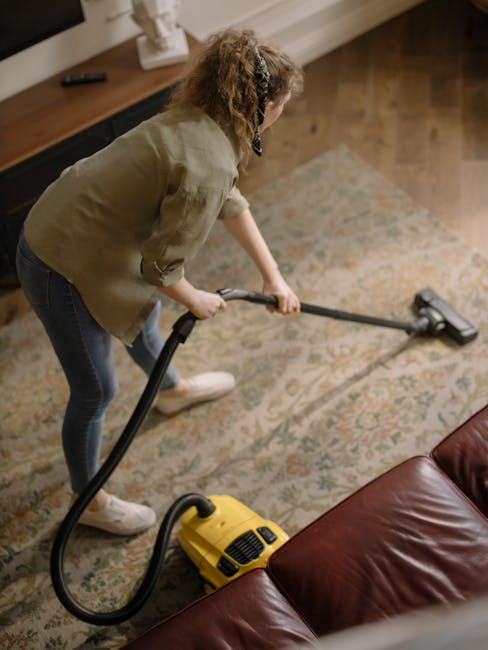A person using a yellow vacuum cleaner equipped with a long black hose and a vacuum head attachment is performing surface cleaning on a patterned area rug in a domestic living room. The rug has a trad