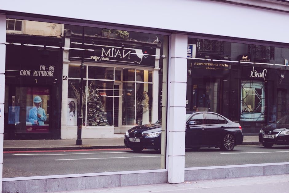 A large glass storefront window reflecting the street scene outside, including parked black cars, a lamppost, and nearby shops with their signage visible. The interior of the shop is not visible throu